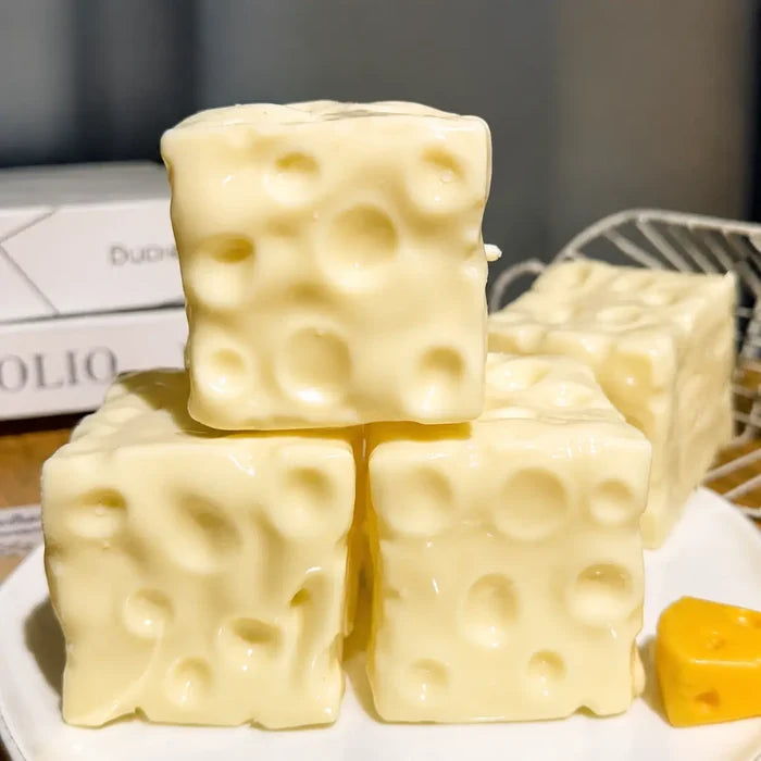 Three pale yellow cheese-shaped blocks stacked on a white plate, with a small yellow cheese slice in the corner and a blurred background.