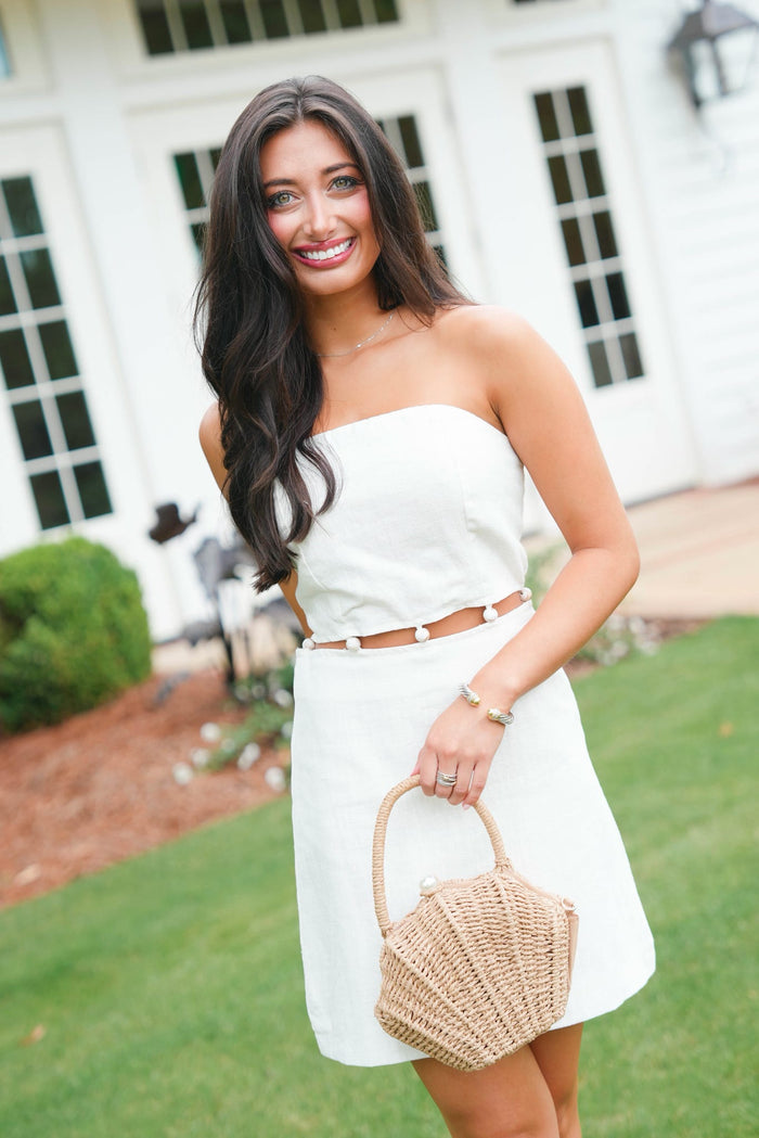 Woman in a white strapless dress holding a woven handbag outdoors.