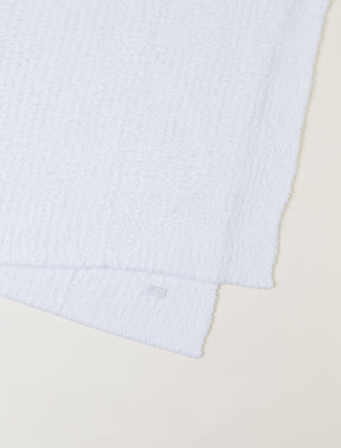 A close-up of a white textured blanket, folded diagonally on a light beige background.
