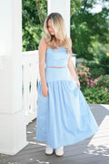 Woman wearing a light blue polka dot dress standing on a wooden deck with greenery in the background