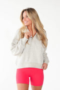 A woman modeling a light heather gray quarter-zip pullover sweatshirt and bright pink athletic shorts against a plain white studio background.