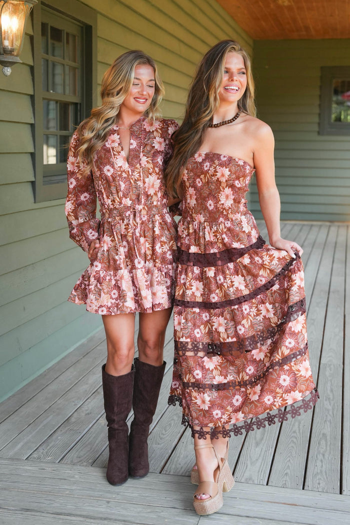 Two women standing on a wooden porch wearing matching floral midi dresses in a dusty rose and brown colorway, with one wearing knee-high brown boots.