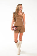 A woman modeling a matching brown two-piece set consisting of a sleeveless top and shorts, paired with beige cowboy boots and holding a football, against a plain white studio background.