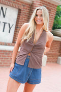A smiling blonde woman wearing a sleeveless brown wrap-style top with a twisted front detail and a side slit, paired with denim shorts, standing outdoors in front of a brick building.