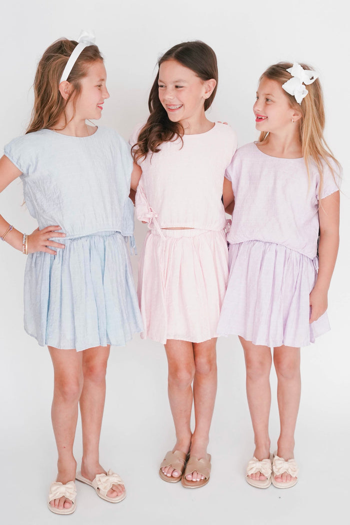 Three young girls wearing light blue, pink, and white dresses standing against a white background.