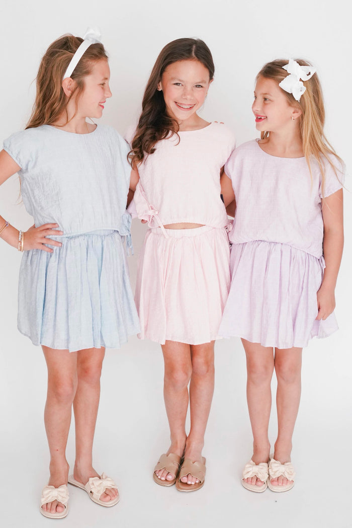 Three young girls wearing light blue, pink, and white dresses standing against a plain background.