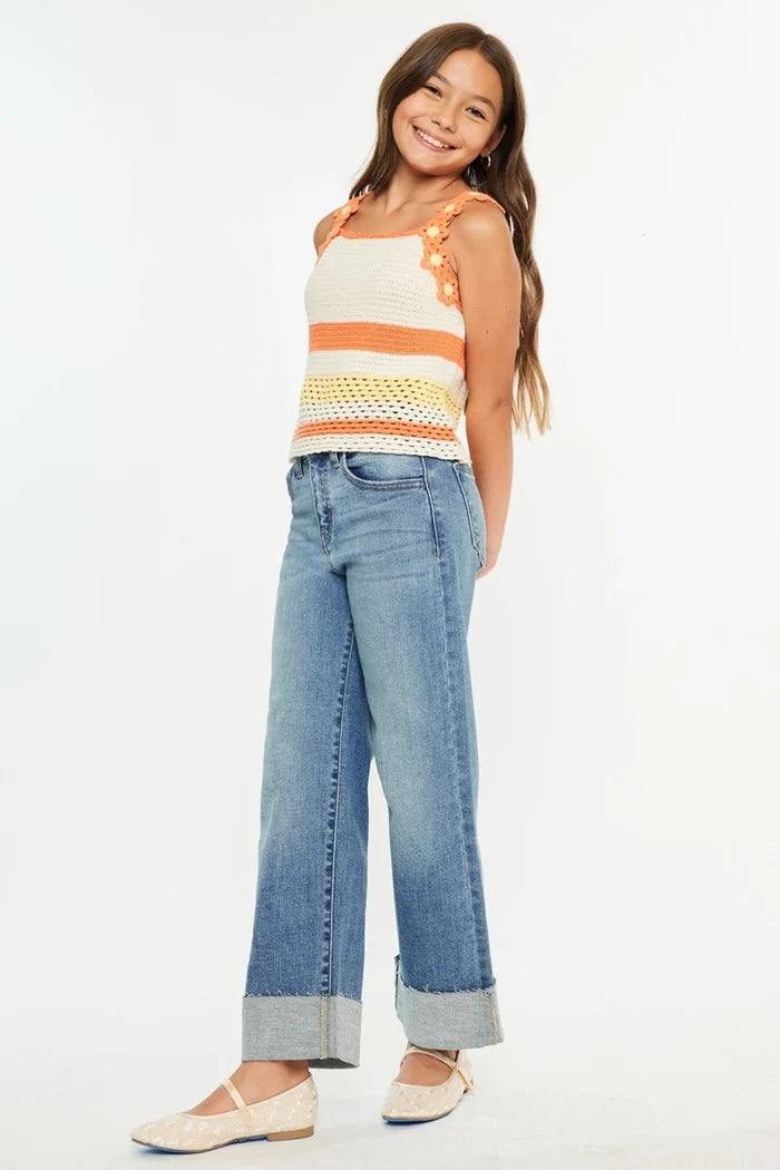 A young girl modeling a sleeveless knit top with orange and cream stripes, blue wide-leg jeans with rolled cuffs, and cream flat shoes against a plain white background.
