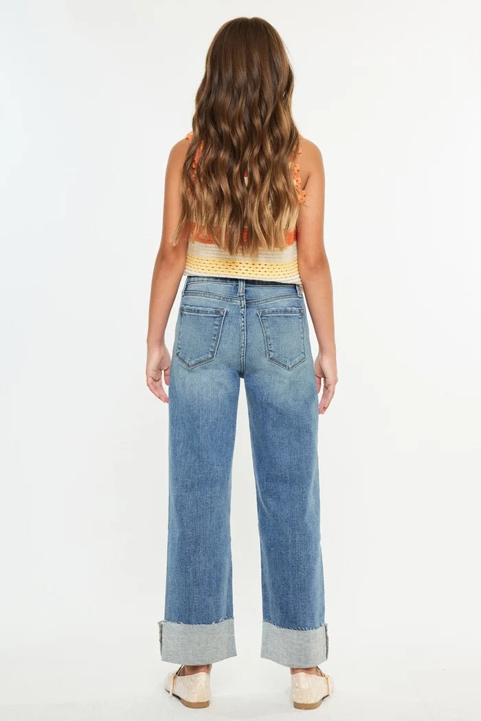 A woman viewed from behind wearing light-wash blue wide-leg jeans with rolled cuffs, paired with a yellow and orange striped knit top and beige flat shoes, against a plain white studio background.