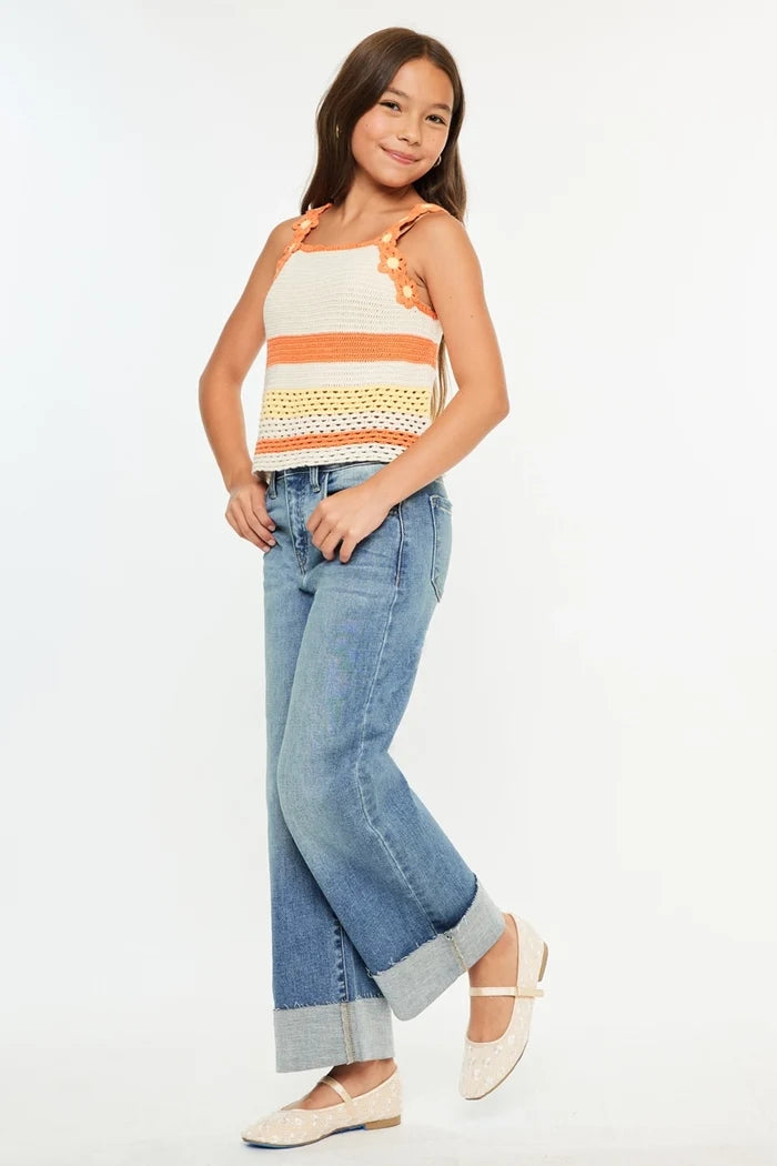 A young girl modeling a sleeveless knit top with orange and yellow stripes, paired with light blue wide-leg jeans and cream flat shoes, against a plain white background.