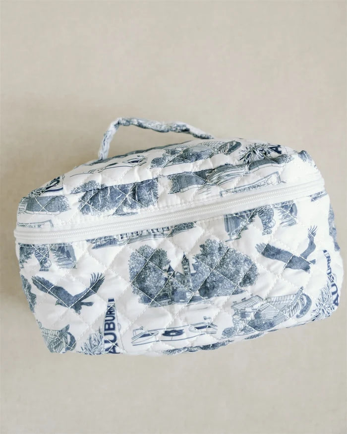 A quilted cosmetic bag with a blue and white toile de Jouy pattern, featuring a top zipper and a matching fabric handle, set against a plain beige background.