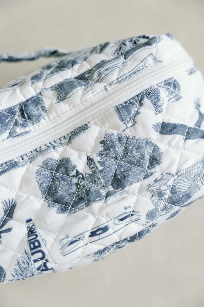 A quilted cosmetic bag with a blue and white toile de Jouy pattern, featuring a top zipper and a matching fabric handle, set against a plain beige background.