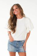 A woman with long wavy brown hair wearing a white puff-sleeve top and a light blue denim mini skirt, posing against a solid white studio background.