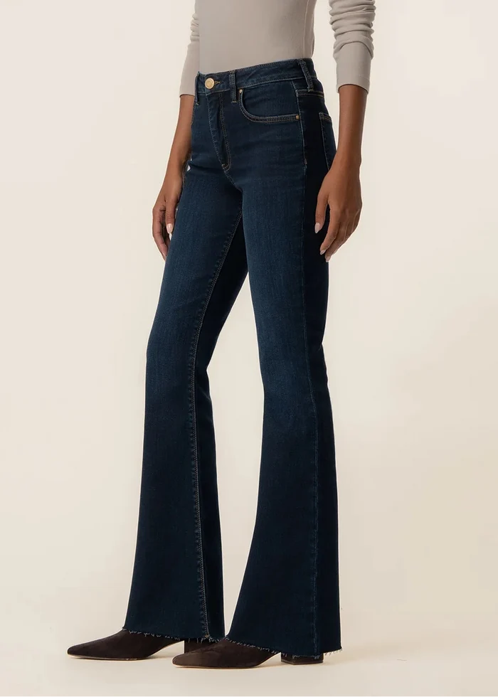 A model wearing dark blue high-waisted flare jeans with a beige long-sleeve top, standing against a plain beige studio background.