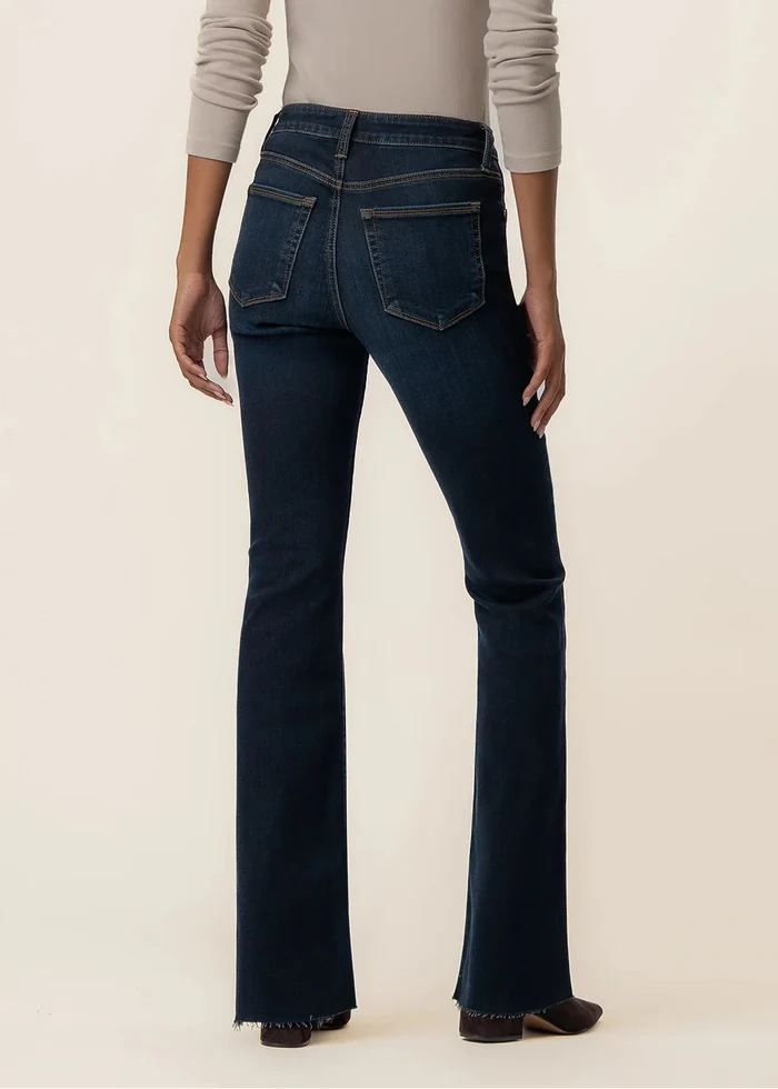 A model shown from the back wearing dark blue flared jeans and a light beige long-sleeve top, standing against a solid cream background.
