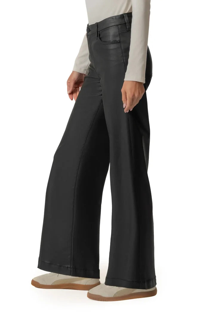 A model wearing black wide-leg pants with a faux-leather finish, styled with a cream long-sleeve top and beige slip-on shoes against a white background.