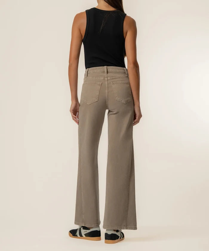 A model shown from the back wearing high-waisted, wide-leg taupe pants, a black sleeveless top, and black-and-white sneakers against a plain off-white studio background.