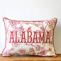 A rectangular decorative throw pillow with a cream fabric base and red floral pattern, featuring the word 'ALABAMA' in large red letters across the front, resting on a wooden surface against a white wall.