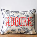 A rectangular decorative throw pillow with a navy blue floral vine pattern on a cream background, featuring the word 'AUBURN' in large red letters across the front, resting on a wooden surface against a light gray wall.