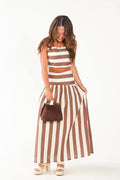 A woman modeling a brown and cream striped two-piece set consisting of a cropped tank top and a matching maxi skirt, holding a small brown handbag against a plain white background.