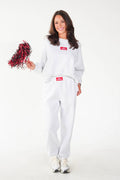 A woman modeling a white sweatshirt and sweatpants set with red Supreme box logos, holding a red and black pom-pom against a plain white background.
