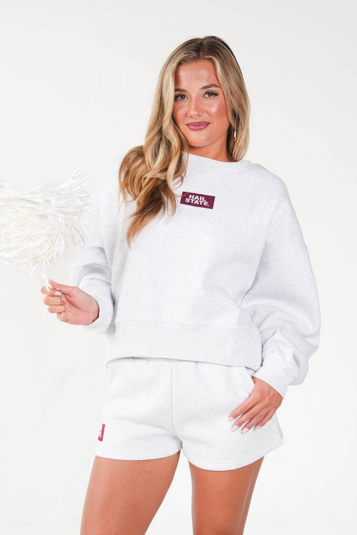 A woman modeling a white crewneck sweatshirt and matching white shorts against a plain white studio background. The sweatshirt features a small maroon rectangular chest patch with white text, and the shorts have a small maroon logo near the left leg.