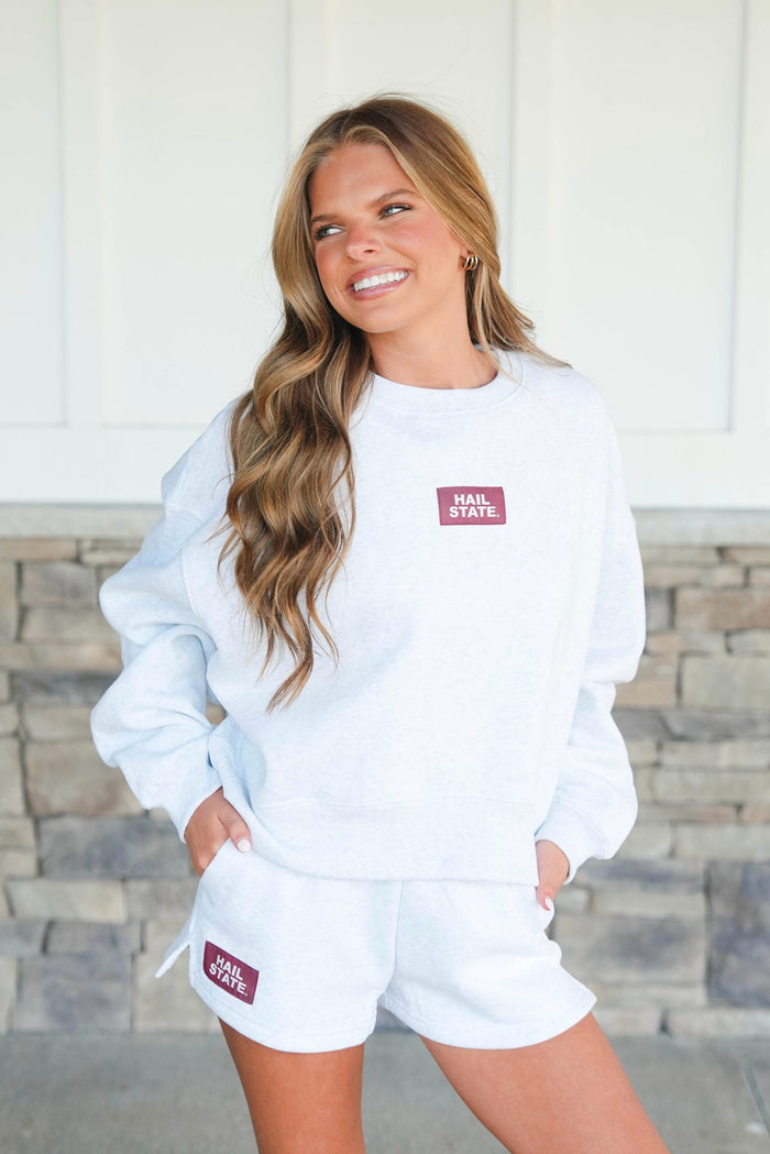 Woman wearing a white sweatshirt and shorts set with a visible brand logo, standing against a stone wall.