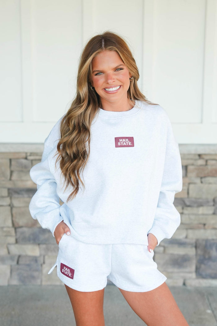 Woman wearing a white sweatshirt and shorts set with a logo, standing against a stone wall.