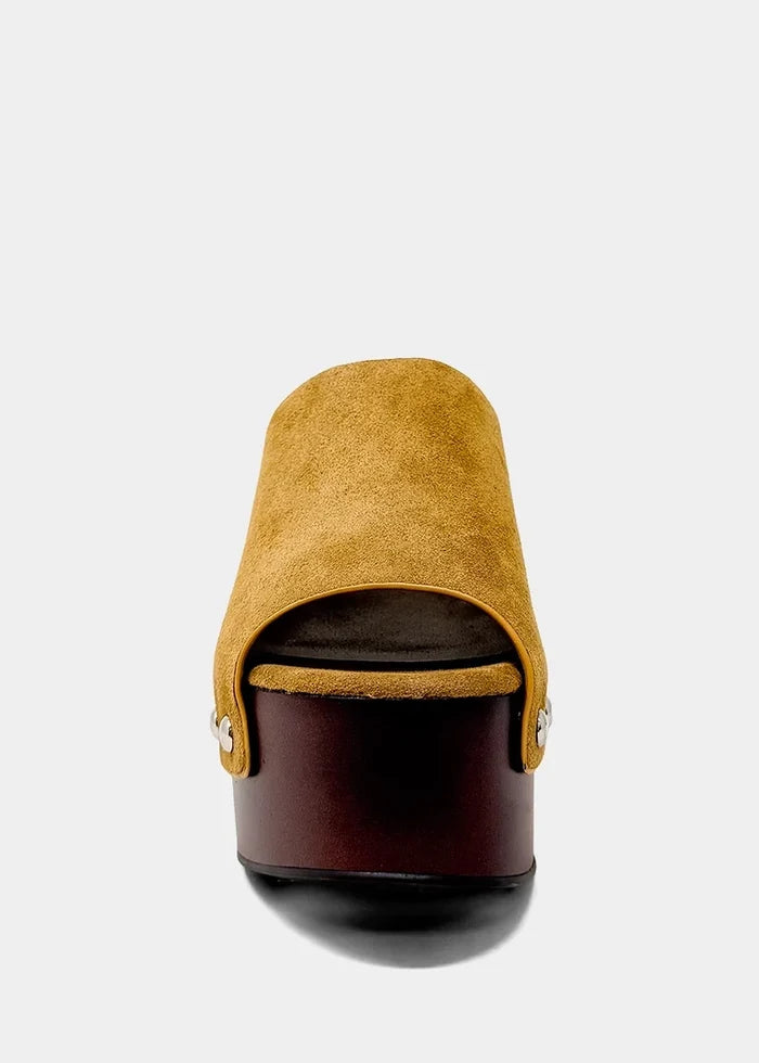 A single platform mule sandal shown from a rear three-quarter view, featuring a mustard yellow suede upper and a dark brown platform sole with a black outsole, set against a light gray background.