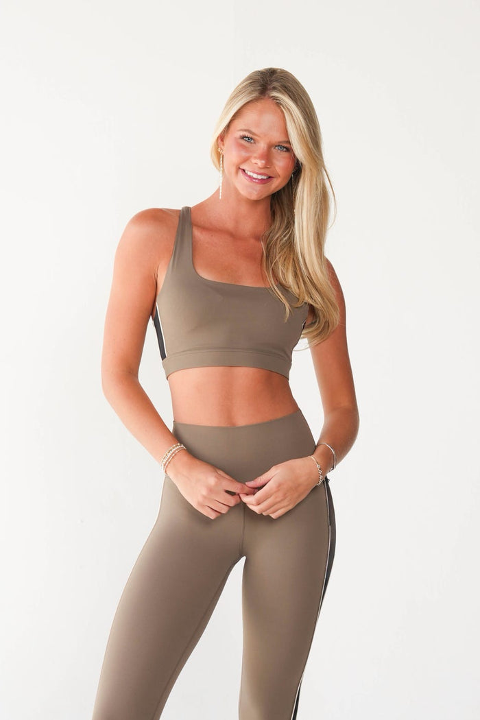 A woman modeling a matching taupe-brown activewear set consisting of a scoop-neck sports bra and high-waisted leggings with a black side stripe, paired with white sneakers against a plain white studio background.