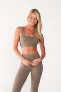 A woman modeling a matching taupe-brown activewear set consisting of a scoop-neck sports bra and high-waisted leggings with a black side stripe, paired with white sneakers against a plain white studio background.