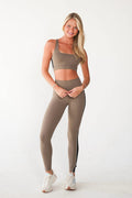 A woman modeling a matching taupe-brown activewear set consisting of a scoop-neck sports bra and high-waisted leggings with a black side stripe, paired with white sneakers against a plain white studio background.
