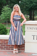A woman stands outdoors in front of a brick wall and a stone pillar, wearing a strapless midi dress with vertical red, white, and navy blue stripes. The background shows green trees and a university-style stone wall with brick paving.