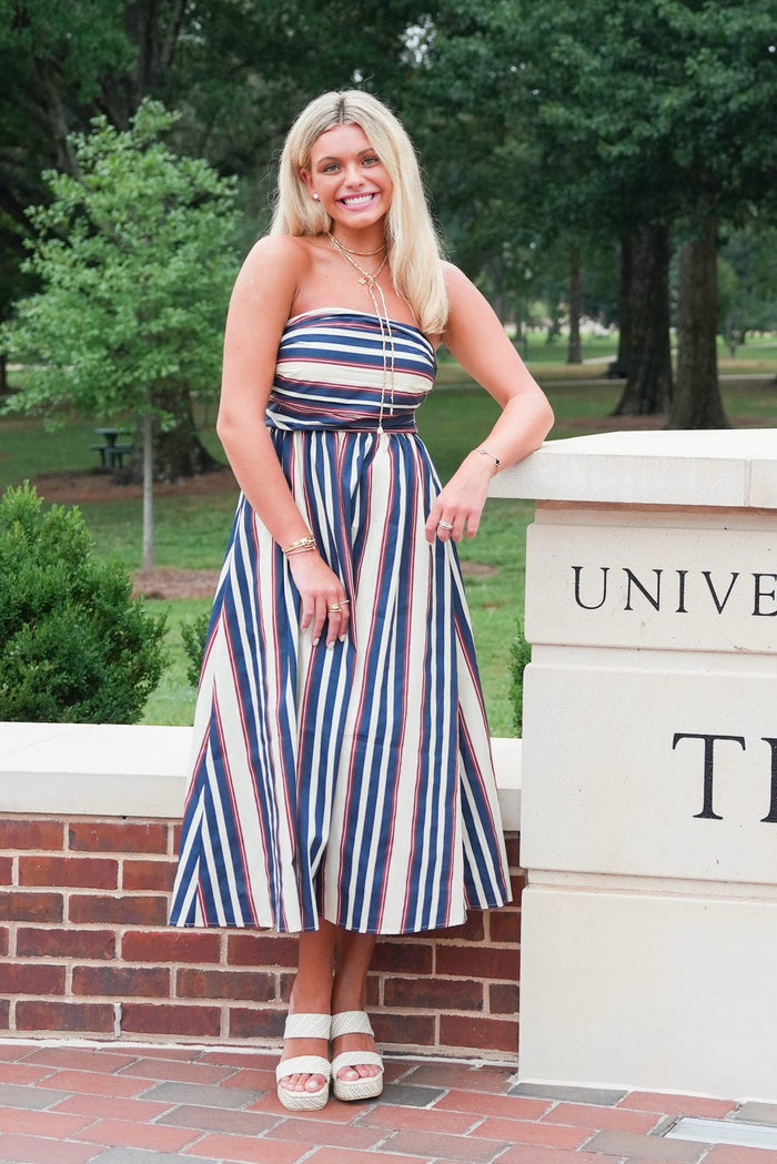 A woman stands outdoors in front of a brick wall and a stone pillar, wearing a strapless midi dress with vertical red, white, and navy blue stripes. The background shows green trees and a university-style stone wall with brick paving.