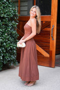A woman wearing a rust-brown strapless maxi dress stands in front of a rustic wooden door, holding a small white clutch.