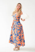 A woman modeling a two-piece floral set consisting of a strapless crop top and a matching high-waisted maxi skirt. The fabric features a vibrant blue base with a bold orange floral pattern.