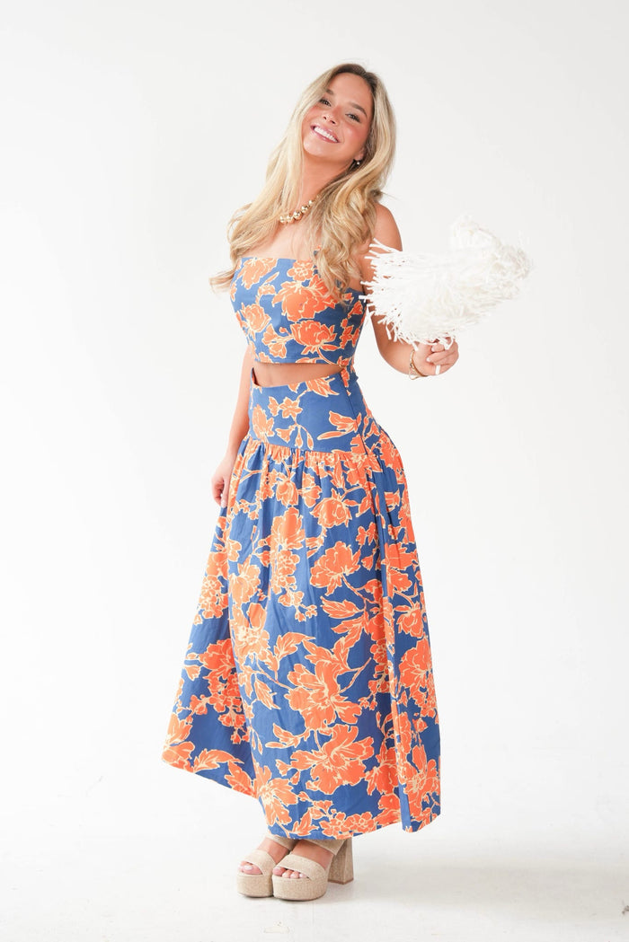 A woman modeling a two-piece crop top and maxi skirt set with a vibrant blue and orange floral pattern, holding a white feather fan against a plain white studio background.