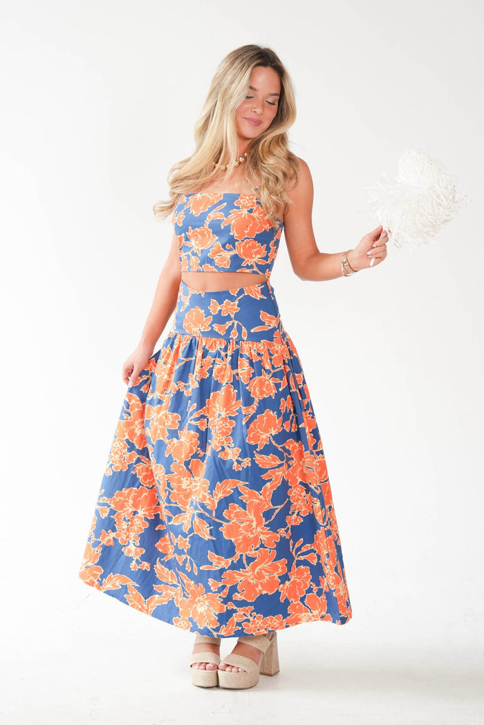 A woman modeling a two-piece halter crop top and matching maxi skirt set with a vibrant blue and orange floral pattern, holding a white feather fan against a plain white studio background.