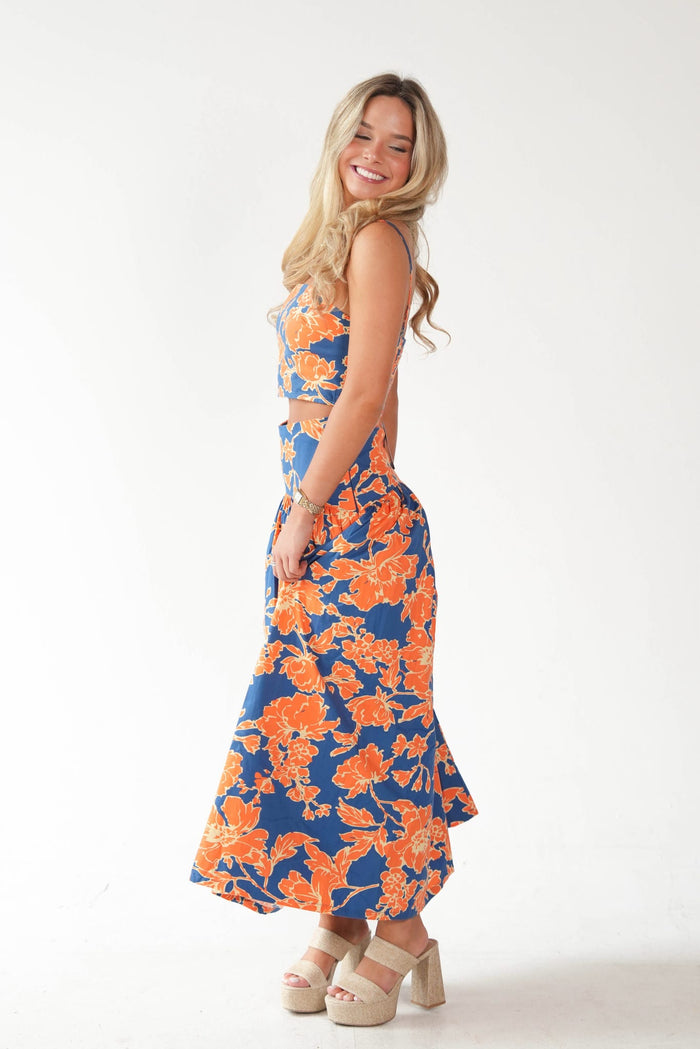 A woman modeling a two-piece sleeveless maxi set with a navy blue base and vibrant orange floral pattern, paired with beige platform sandals, against a plain white studio background.