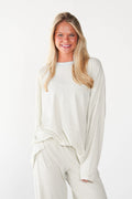 A smiling blonde woman wearing a matching cream-colored ribbed knit lounge set, featuring a long-sleeve top and wide-leg pants, against a plain white studio background.